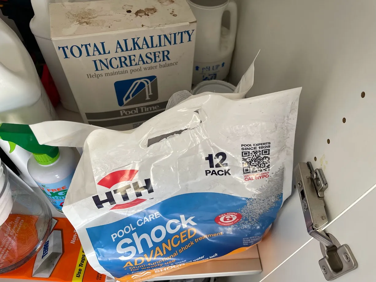 The pool cabinet partway through my DIY years. The alkalinity increaser and pH Up came with the house. The bag of pool shock up front is the one thing I added myself, while flailing, before I understood my real problem was a sky-high CYA level.